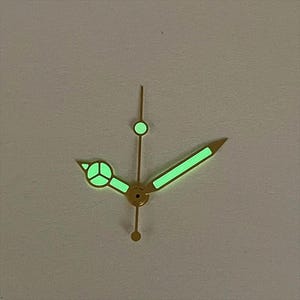 May include: Close-up of watch hands with a gold finish. The hands are illuminated with a bright green glow. The hour hand has a circular design, and the minute hand is a long, straight line. The second hand is a thin line with a circular tip.