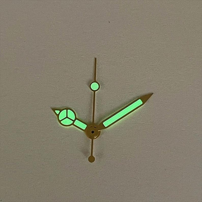 May include: Close-up of watch hands with a gold finish. The hands are illuminated with a bright green glow. The hour hand has a circular design, and the minute hand is a long, straight line. The second hand is a thin line with a circular tip.