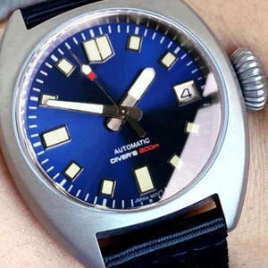 K&ouml;nnte beinhalten: Silberfarbene Uhr mit tiefblauem Zifferblatt und schwarzem Armband. Das Zifferblatt hat wei&szlig;e Zeiger und Markierungen, eine Datumsanzeige und den Text "AUTOMATIC DIVER'S 200m". Die Uhr hat eine strukturierte Krone und ein schwarzes Stoffarmband.
