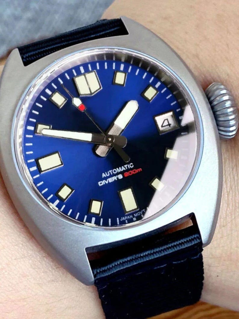 K&ouml;nnte beinhalten: Silberfarbene Uhr mit tiefblauem Zifferblatt und schwarzem Armband. Das Zifferblatt hat wei&szlig;e Zeiger und Markierungen, eine Datumsanzeige und den Text "AUTOMATIC DIVER'S 200m". Die Uhr hat eine strukturierte Krone und ein schwarzes Stoffarmband.