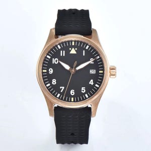 May include: A bronze-colored wristwatch with a black face and white numerals. The watch has a black rubber strap and a date display at the 3 o'clock position. The watch case is approximately 40mm in diameter.