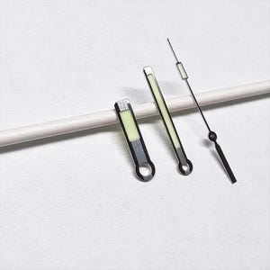 May include: A set of three watch hands. Two are rectangular with a luminous green center and silver trim. The third is a thin, black hand with a small circle and a rectangular luminous green tip. The hands are for a watch.