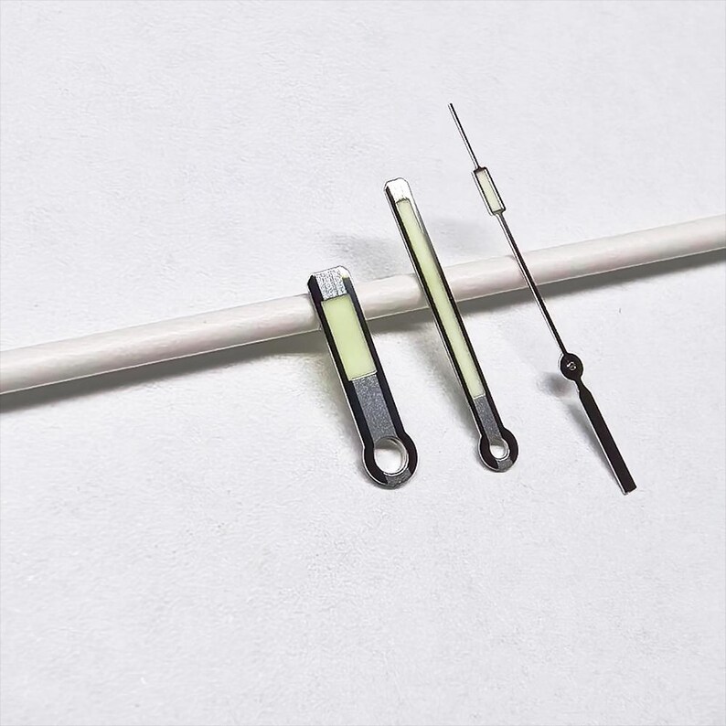 May include: A set of three watch hands. Two are rectangular with a luminous green center and silver trim. The third is a thin, black hand with a small circle and a rectangular luminous green tip. The hands are for a watch.