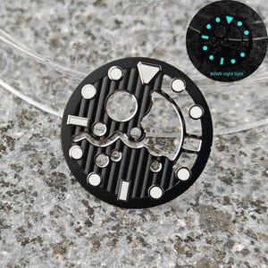 May include: Black watch dial with silver and white accents. The dial features a circular design with a ribbed texture and various geometric shapes. A smaller, glowing version of the dial is shown, with the text "BGW9 night light".
