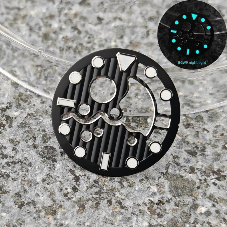 May include: Black watch dial with silver and white accents. The dial features a circular design with a ribbed texture and various geometric shapes. A smaller, glowing version of the dial is shown, with the text "BGW9 night light".