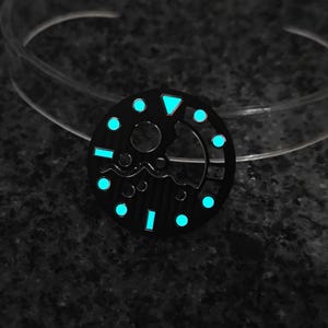 May include: A black watch dial with luminous blue markers and a triangle at the 12 o'clock position. The dial has a circular design with a central opening and a wave-like pattern. The markers and hands glow in the dark.