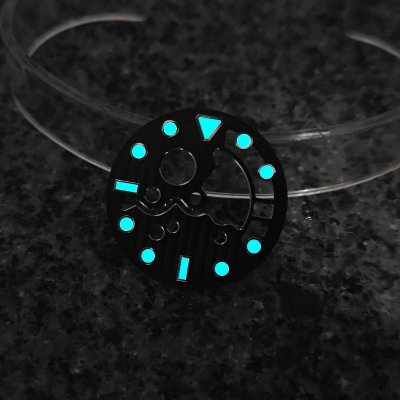 May include: A black watch dial with luminous blue markers and a triangle at the 12 o'clock position. The dial has a circular design with a central opening and a wave-like pattern. The markers and hands glow in the dark.