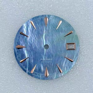 Puede incluir: Una esfera de reloj con una superficie azul nacarada, con marcadores de hora y agujas de color oro rosa. El texto "PRESAGE AUTOMATIC" est&aacute; impreso en la esfera. La esfera del reloj tiene una ventana de fecha y est&aacute; marcada "MADE IN JAPAN".