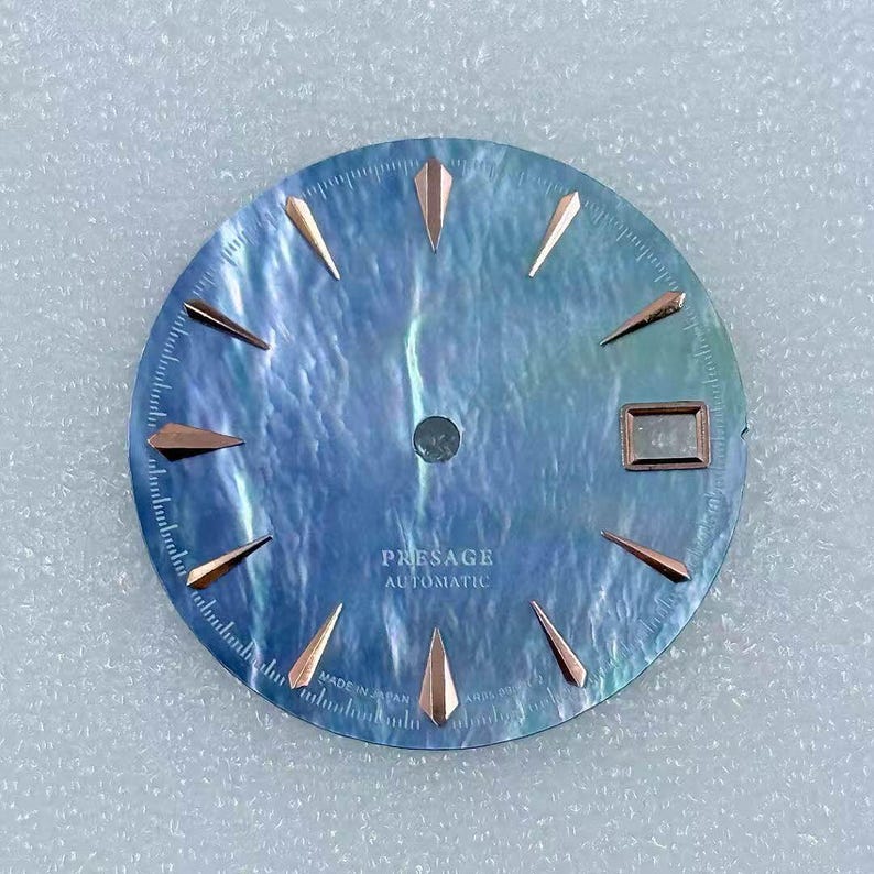 Puede incluir: Una esfera de reloj con una superficie azul nacarada, con marcadores de hora y agujas de color oro rosa. El texto "PRESAGE AUTOMATIC" est&aacute; impreso en la esfera. La esfera del reloj tiene una ventana de fecha y est&aacute; marcada "MADE IN JAPAN".