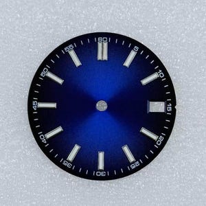 May include: A watch dial with a blue gradient face. It has white rectangular hour markers and a minute track around the edge. The numbers 5, 10, 15, 20, 25, 30, 35, 40, 45, 50, 55, and 60 are printed in white.