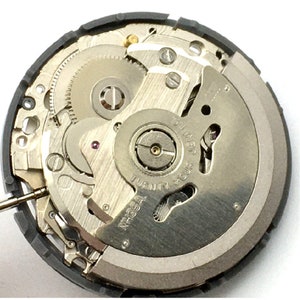 K&ouml;nnte beinhalten: Nahaufnahme eines silbernen Uhrwerks mit dem sichtbaren Text "NH35A TWENTY-FOUR JEWELS". Das Uhrwerk ist zerlegt und zeigt das Innenleben der Uhr.