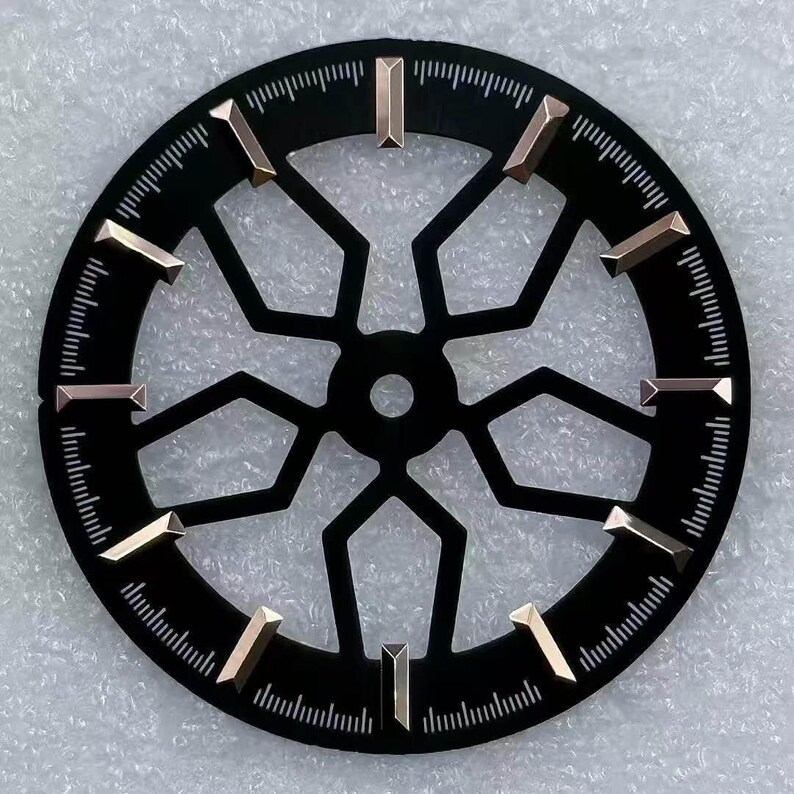 Pode incluir: Um mostrador de rel&oacute;gio preto com um design geom&eacute;trico e marcadores de hora dourados. O mostrador tem formato circular com marca&ccedil;&otilde;es de minutos na borda. Os marcadores de hora s&atilde;o retangulares e angulados.