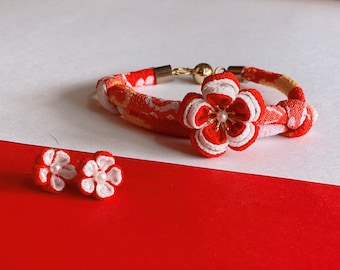 May include: A red and white fabric bracelet with a flower charm and matching red and white fabric earrings. The bracelet has a gold clasp and a pearl in the center of the flower charm.