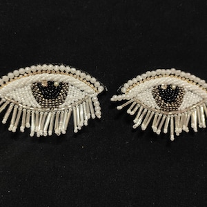 May include: Two white beaded eye patches with black and silver accents. The eyes have a fringe of silver beads around the edges.