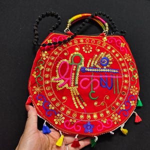 Puede incluir: Un bolso rojo bordado con un dise&ntilde;o circular con lentejuelas doradas e hilo de colores. El bolso tiene una correa de cuentas y un asa de color arco&iacute;ris. Borlas de varios colores cuelgan de la parte inferior.