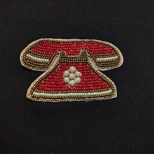 Op de afbeelding: Een rode, met kralen bezette telefoonpatch met gouden en witte accenten. De patch heeft de vorm van een vintage telefoon, met een bloemachtig ontwerp in het midden. Op een zwarte achtergrond.