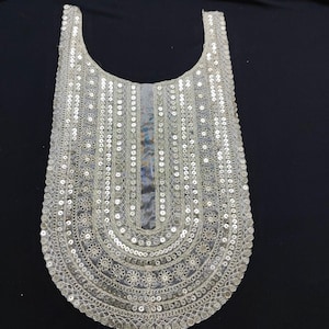 May include: A silver-toned, decorative collar or neckline element. It is densely adorned with small, circular sequins, organized in vertical and curved designs. The piece has a rounded lower edge and a gently curved upper edge, indicating it's designed to be worn around the neck.