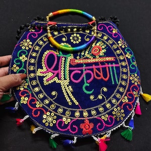 May include: A navy blue, embroidered handbag with a rainbow handle. The bag features colorful embroidery, sequins, and tassels. The design includes text in a script font. The bag is held by a hand.