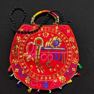 Puede incluir: Un bolso rojo bordado con un dise&ntilde;o circular que presenta texto colorido y una pluma de pavo real. El bolso tiene una correa de cuentas y un asa de arco&iacute;ris. Lentejuelas doradas y borlas coloridas adornan los bordes.