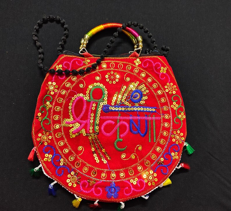 Puede incluir: Un bolso rojo bordado con un dise&ntilde;o circular que presenta texto colorido y una pluma de pavo real. El bolso tiene una correa de cuentas y un asa de arco&iacute;ris. Lentejuelas doradas y borlas coloridas adornan los bordes.