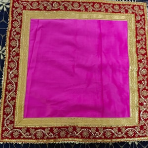 May include: A square textile with a bright fuchsia centre. The border is red, with gold-coloured embroidery and sequins. The design features floral and scroll patterns, creating an ornate and elegant look. The textile is likely used for decorative purposes.