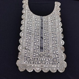 May include: A silver-colored, intricately embroidered collar with a scalloped edge. The collar features a floral pattern and is embellished with sequins. The design is symmetrical and appears to be made of a delicate, sheer fabric.
