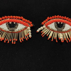 May include: A pair of beaded eye earrings with orange, white, black and gold beads. The earrings have a fringe of gold beads.
