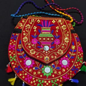 May include: A round red fabric bag featuring intricate multi-colored embroidery, small circular mirrors, and gold sequins. The bag has a flap closure, a zipper, and a braided multi-colored strap, with small tassels hanging from the bottom edge.