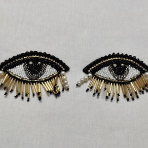 Könnte beinhalten: Zwei schwarze und weiße Perlen-Augenklappen mit goldenen Fransen und weißen Perlenakzenten.