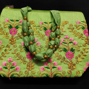 Può includere: Una borsa a mano in tessuto verde con una tracolla a perline. La borsa è decorata con motivi floreali ricamati in rosa e oro.