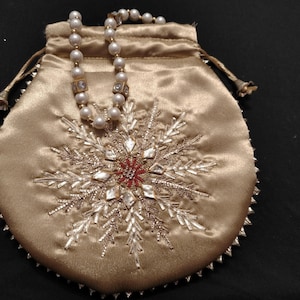 Puede incluir: Una bolsa de cordón de satén dorado con un diseño de copo de nieve bordado con cuentas plateadas y blancas. Un collar de perlas con detalles dorados está dentro de la bolsa.