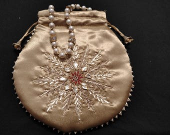 Puede incluir: Una bolsa de cordón de satén dorado con un diseño de copo de nieve bordado con cuentas plateadas y blancas. Un collar de perlas con detalles dorados está dentro de la bolsa.