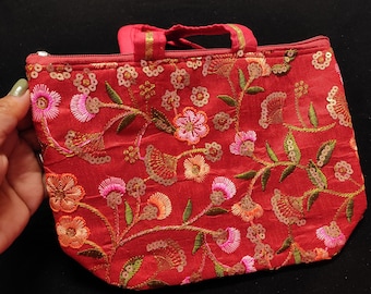 Puede incluir: Un pequeño bolso rojo con un diseño de bordado floral. El bolso tiene un cierre de cremallera y un asa superior.