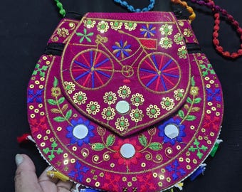 Puede incluir: Un bolso bandolera circular de color magenta con un motivo de bicicleta bordado en la solapa. El bolso está decorado con lentejuelas doradas, pequeños espejos circulares y bordados florales coloridos. Incluye una correa de cuentas multicolor.