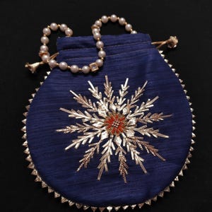 Puede incluir: Una bolsa de cordón de seda azul con un diseño de copo de nieve dorado y un collar de perlas. La bolsa es redonda y tiene un borde festoneado.