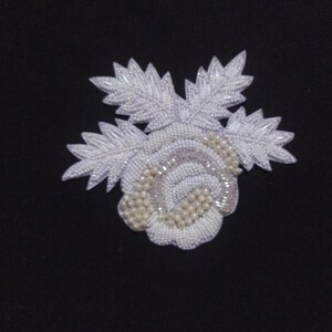 Pode incluir: Uma aplicação de flor branca com contas com um design de rosa e folhas. A flor é feita de pequenas contas e pérolas brancas.
