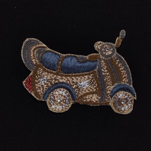 May include: A beaded applique of a gold and silver scooter with a blue seat. The scooter has a red tail light and is on a black background.