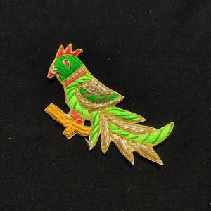 May include: A green and gold embroidered bird applique with a long tail. The bird is perched on a branch with yellow beads.