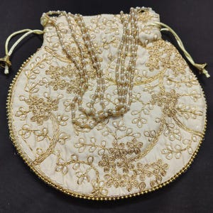 Pode incluir: Uma bolsa redonda de cor creme com fecho de cordão, apresentando um intrincado bordado de fio dourado com padrões florais e de videiras. A bolsa é acentuada com uma alça de pérolas e uma borda delicada de pequenas contas douradas.