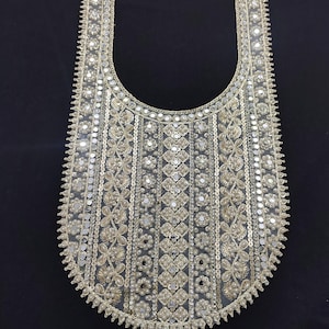 May include: A silver and white embroidered neckline with intricate floral and geometric patterns. The embellishments include sequins and beads, creating a shimmering effect. The neckline is set against a black background, highlighting the detailed design.