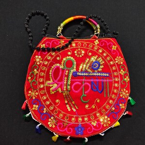 Puede incluir: Un bolso rojo bordado con un dise&ntilde;o circular que presenta texto colorido y adornos. El bolso tiene una correa de cuentas negras y un asa con detalles de arco&iacute;ris. Peque&ntilde;as borlas de varios colores adornan el borde inferior.