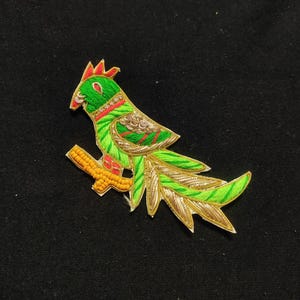 Może przedstawiać: Naszywka z haftowanego ptaka w kolorze zielonym i złotym z długim ogonem. Ptak siedzi na gałęzi ozdobionej żółtymi koralikami. Ta naszywka idealnie nadaje się do dodania odrobiny fantazji do ubrań lub rękodzieła.
