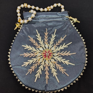 Könnte beinhalten: Eine graue Satin-Kordeltasche mit goldfarbener Perlenborte und einem gestickten Schneeflockenmuster in Gold- und Silberfaden. Die Schneeflocke hat eine rote und weiße Mitte.