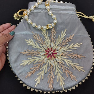Pode incluir: Uma bolsa de cordão de veludo cinza com um desenho de floco de neve bordado em fio dourado e prateado. A bolsa tem uma corrente de contas de pérolas e cristais presa ao topo.