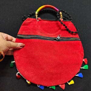 Puede incluir: Un bolso de mano redondo rojo con cremallera negra y borlas coloridas. El asa est&aacute; hecha de cuentas negras y una secci&oacute;n envuelta de colores del arco&iacute;ris. El bolso es sostenido por una mano con una pulsera dorada.