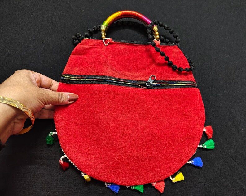 Puede incluir: Un bolso de mano redondo rojo con cremallera negra y borlas coloridas. El asa est&aacute; hecha de cuentas negras y una secci&oacute;n envuelta de colores del arco&iacute;ris. El bolso es sostenido por una mano con una pulsera dorada.