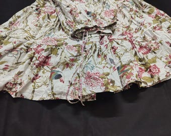 May include: A white tiered skirt featuring a botanical print with pink flowers, green leaves, and colorful birds. The skirt has an elasticized waistband with a drawstring tie, creating a gathered, voluminous silhouette with multiple ruffled layers.