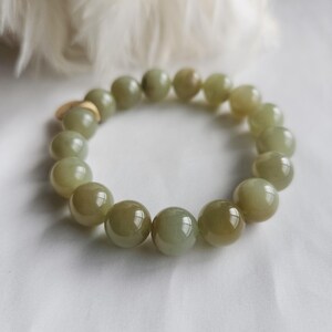 May include: A bracelet composed of large, spherical, olive-green beads. The beads are polished and reflective. The bracelet is presented on a white surface, with a soft, white, feathery texture in the background. The beads are approximately 1.5 cm in diameter.