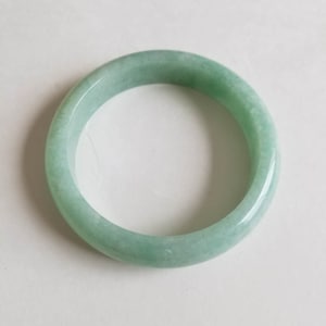 May include: A circular jadeite bangle in a pale green hue. The polished stone has a smooth, glossy finish and a translucent quality. The bracelet is a complete, unbroken loop, displaying the natural colour variations of the jade.