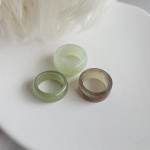 May include: Three smooth, solid band rings with a polished finish. One ring is a pale, translucent mint green, another is a deeper sage green, and the third is a muted, semi-transparent brownish-gray. Each ring features a simple, rounded profile.